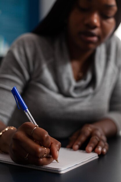 Close up of woman taking notes with pen on textbook at desk. Young adult writing information on notebook papers while working from home on business project. Person with remote work