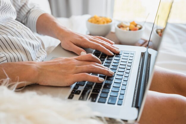 Close-up of woman typing on laptop with breakfast on bed