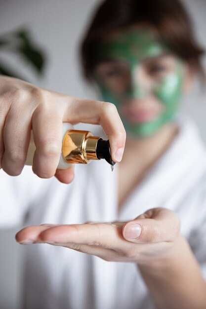 Close-up, a woman in a white robe with a green cosmetic mask presses on the dispenser of beauty care products, blurred background.