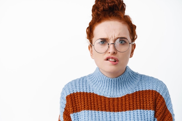 Free photo close up of worried and troubled redhead woman in glasses, frowning and biting lip puzzled, have proble, staring complicated on white