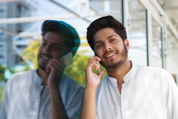 Close-up of young Arabian man holding phone. Handsome bearded man speaking over mobile phone reflecting in shop glass outdoors. Communication and modern technology concept