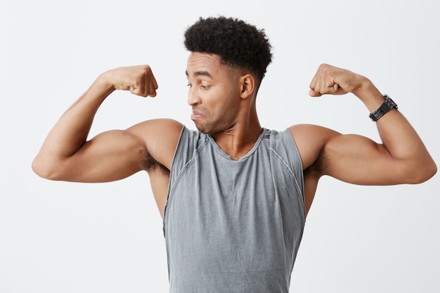 Foto gratuita chiuda in su di giovane uomo dalla pelle scura bello atletico con l'acconciatura afro in camicia grigia sportiva guardando i suoi muscoli con espressione del viso concentrato e fiducioso. uno stile di vita sano