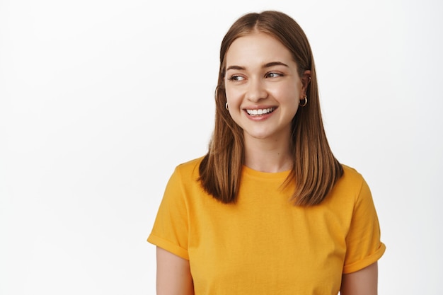 Close up of young attractive woman with fair short hair, wearing yellow t-shirt, smiling white teeth, looking left at empty space for sale banner, logo place, standing over white wall