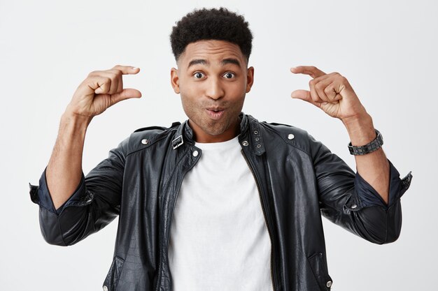 Close up of young beautiful dark-skinned male student with afro hairstyle in white t-shirt under black leather jacket gesticulating with both hands, looking in camera with surprised face expression.