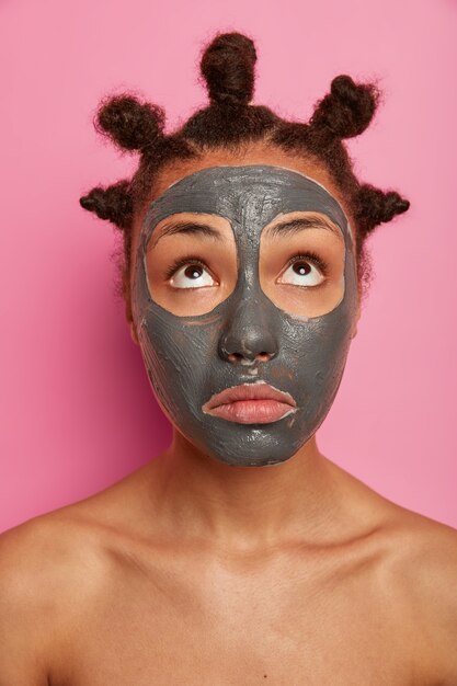 Close up on young beautiful woman with facial maskoman concentrated above, wears facial clay mask for reducing acnes, stands naked, cares about body, isolated on pink background, has many combed buns on head