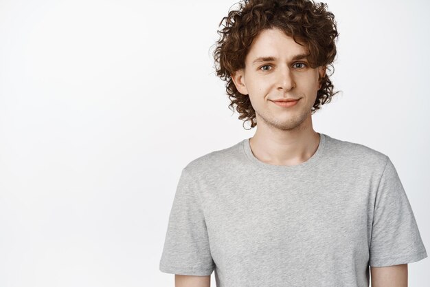 Close up of young caucasian guy with curly hairstyle smiling and looking at camera standing in grey tshirt over white background