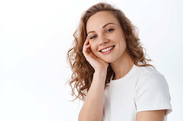Close up of young caucasian natural blond with light make up and happy perfect smile, touching face and look at front, stands against white wall