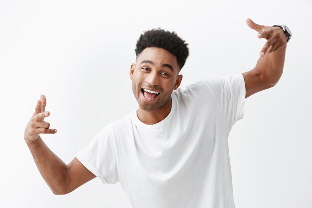 Close up of young cheerful dark-skinned beautiful african man with curly hair in white t-shirt laughing, gesticulating with hand, looking in camera with joyful and happy expression.