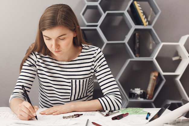 Close up of young concentrated beautiful young designer sitting at table in light room, doing for blueprints using pen and ruler. Business concept