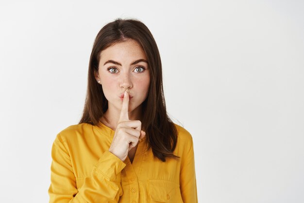 Close-up of young cute woman hushing, telling a secret, holding finger on mouth and looking at camera, standing over white wall