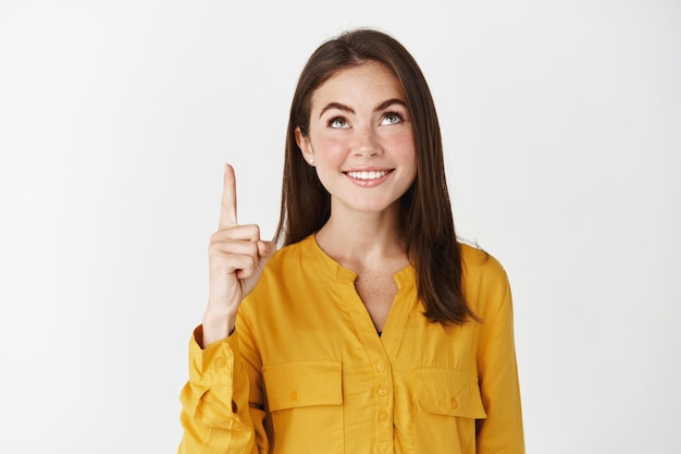 Close-up of young female student smiling, looking and pointing finger up, showing promo offer on copy space, standing over white wall