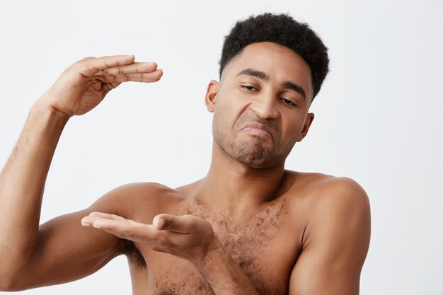 Close up of young funny black-skinned men with curly hair without clothes gesticulating with hands, showing big size, looking aside with cynical expression.