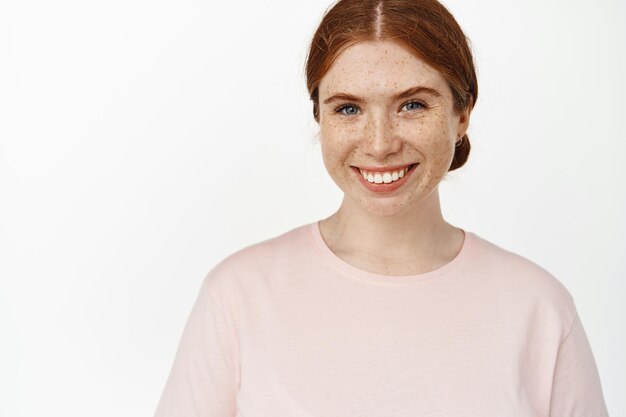 Close up of young ginger girl, caucasian woman with red hair, freckles and white perfect teeth, smiling happy at camera, positive face expression, standing against white background