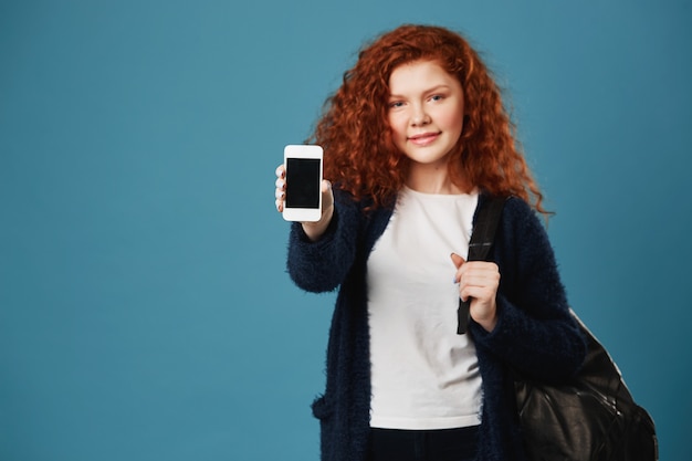 Close up of young ginger student woman with wavy hair and freckles wearing white t-shirt and black cardigan showing smartphone.