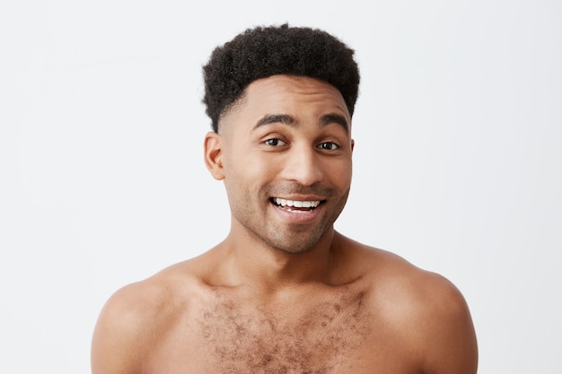 Close up of young good-looking attractive cheerful black-skinned man with afro hairstyle with naked torso smiling with teeth, looking in camera with happy an relaxed expression. Health and beauty