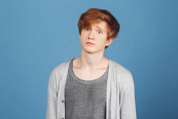 Close up of young good-looking happy red-haired guy in gray t-shirt under cardigan  with confused expression, after receiving hard question on exam in university.