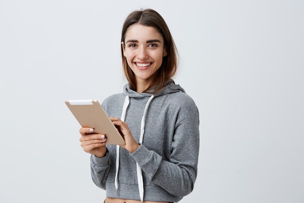 Close up of young good-looking joyful caucasian girl with dark long hair in gray sporty hoodie smiling with teeth, playing games on digital tablet,