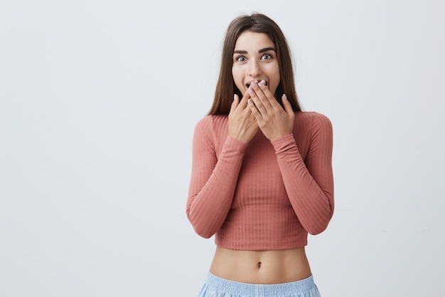 Close up of young handsome cheerful caucasian girl with dark long hair in pink shirt closing mouth with hands with happy and surprised expression