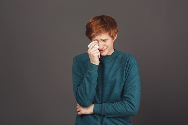 Close up of young handsome sensitive ginger teenager in green sweater crying, wipes tears with paper napkin, being tired of bad relationships with parents, that don't allow him go to the party.