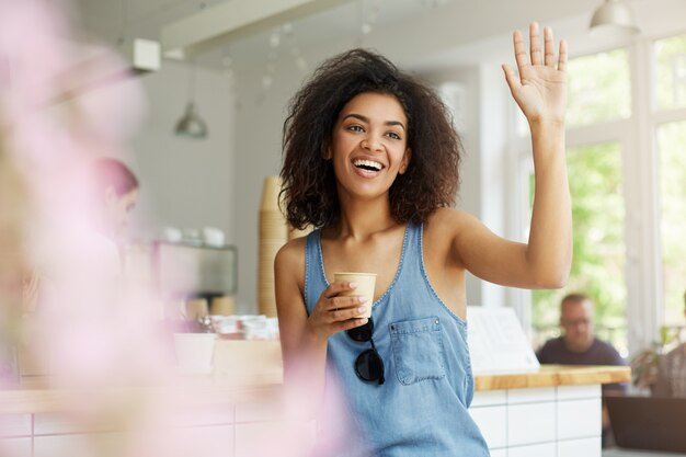 Close up of young joyful dark-skinned student woman with curly dark hair in casual blue shirt sitting in cafeteria, drinking coffee, wagging friend with happy and excited expression.