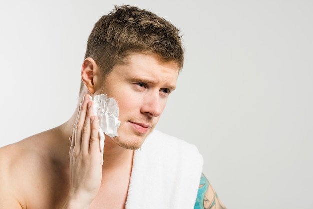 Free photo close-up of a young man applying shaving foam on his cheek against grey background