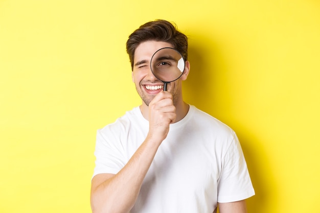 Close-up of young man looking through magnifying glass and smiling, searching something, standing over yellow background.