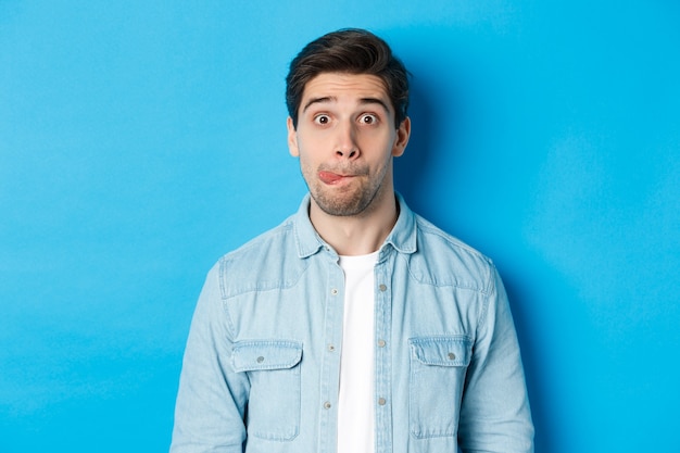 Close-up of young man making funny expressions, showing tongue and looking at camera, standing over blue background