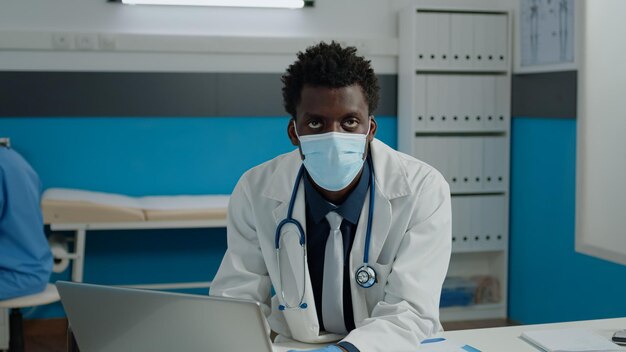 Close up of young medic with face mask in medical cabinet using modern laptop on desk. Doctor sitting with digital device typing on keyboard while wearing stethoscope for healthcare