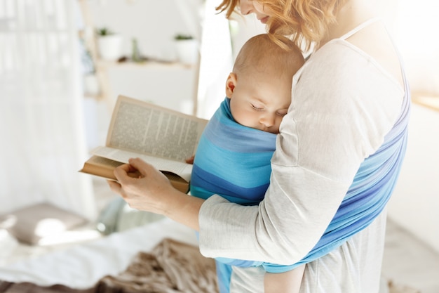 Close up of young mom reading fairy tales for her newborn little son in comfortable light bedroom. Baby falls asleep while she was reading.