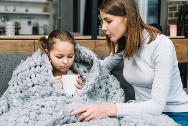 Close-up of young mother taking care of her sick daughter holding mug in hand