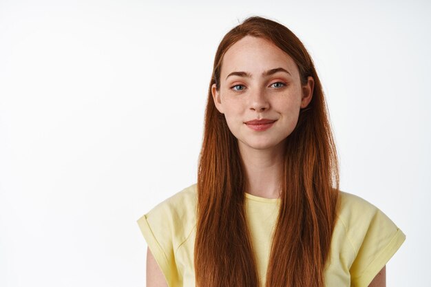 Close up of young motivated girl with red long hair, confident smile and determined attitude, looking at camera, standing against white background.