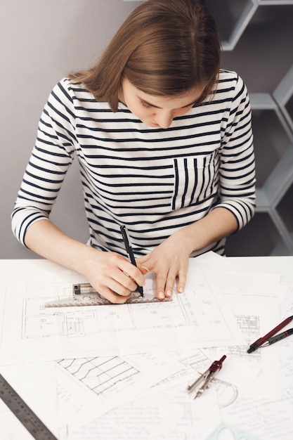 Free photo close up of young professional handsome skinny dark-haired girl in striped shirt, working on new blueprint for future team project