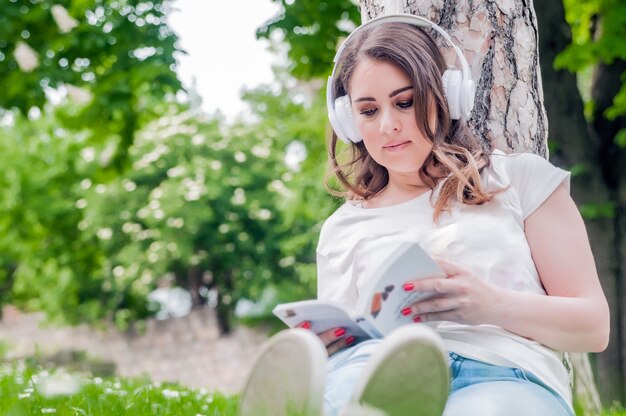 Close-up of young relaxed woman reading and listening music on headphones outside in a spring day. Freetime concept with beautiful female relaxing outdoors with music, books and coffee