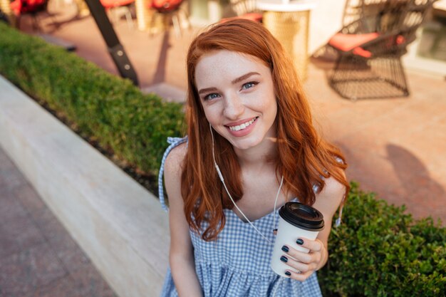 Close up of a young smiling redhead girl in earphones holding cup of coffee