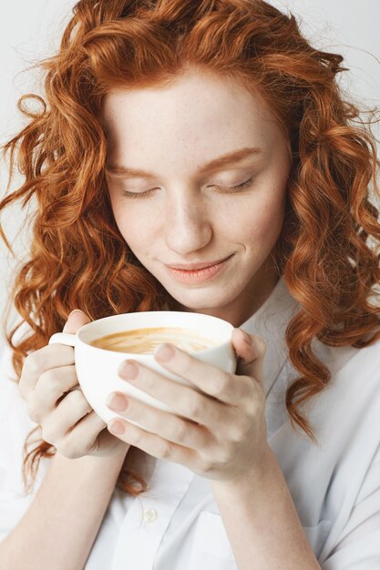 保持している一杯のコーヒーを笑顔で目を閉じて若い優しい赤毛の女の子のクローズアップ。