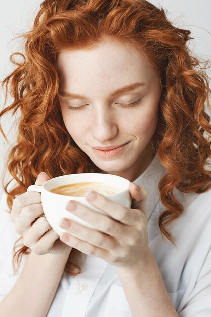 Close up of young tender redhead girl with closed eyes smiling holding cup of coffee.