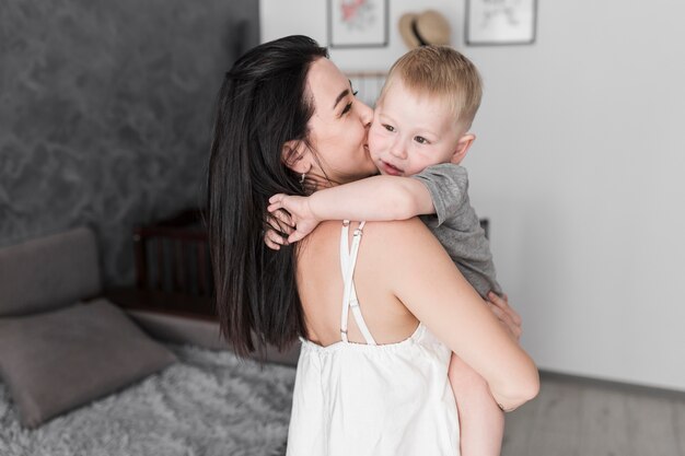 Close-up of young woman carrying her little cute son kissing on his cheeks