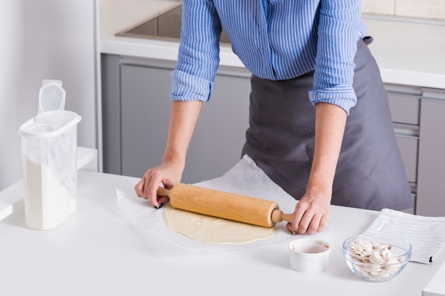 Close-up of young woman flattened dough with rolling pin on white table