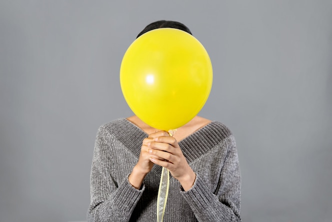 Close up of young woman hiding her face with yellow ballon against gray wall. color trends of 2021. positive concept