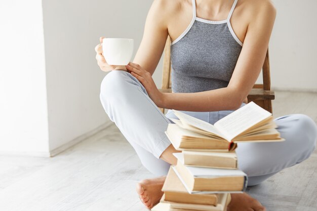 Close up of young woman  holding book cup of coffee sitting on floor with books over white wall early in morning.