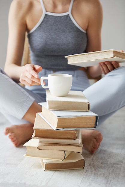 Close up of young woman  holding book and cup sitting on floor over white wall early in morning.