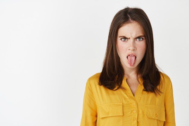 Close-up of young woman showing tongue and disappointment, frowning upset, staring at something disgusting, express dislike while standing over white wall.