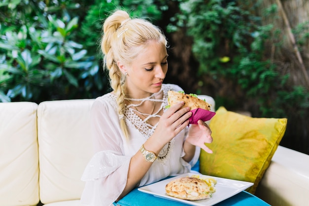 Free photo close-up of young woman sitting on sofa eating sandwich at outdoors