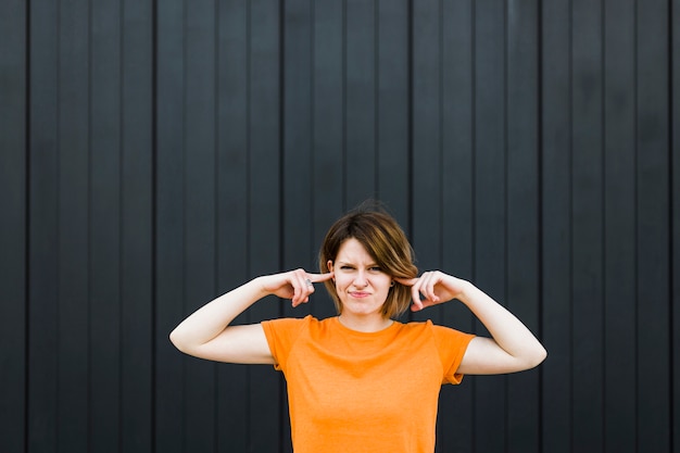 Close-up of a young woman standing against black wall closing her ears with two fingers