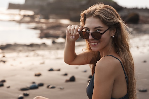 Close-up of a young woman in swimsuit and sunglasses, sit on a deserted tropical beach on black volcanic sand.