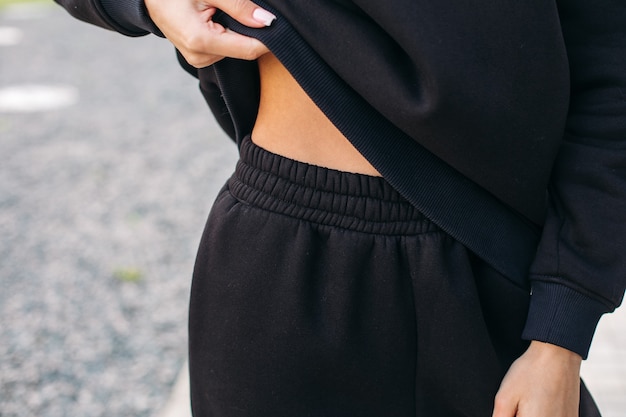 Close up of young woman wearing black tracksuit and lifting the hoodie with her hand. Female fashion. Copy space