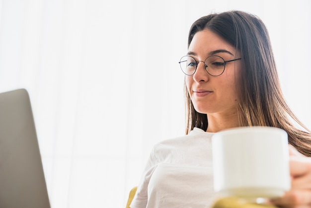 Close-up of young woman wearing eyeglasses holding coffee cup using laptop