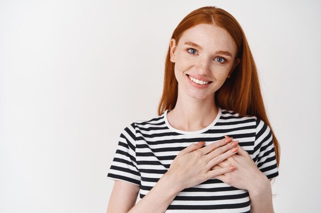 Close-up of young woman with long red hair and blue eyes, thanking you, holding hands on heart and smiling heartfelt, standing over white wall