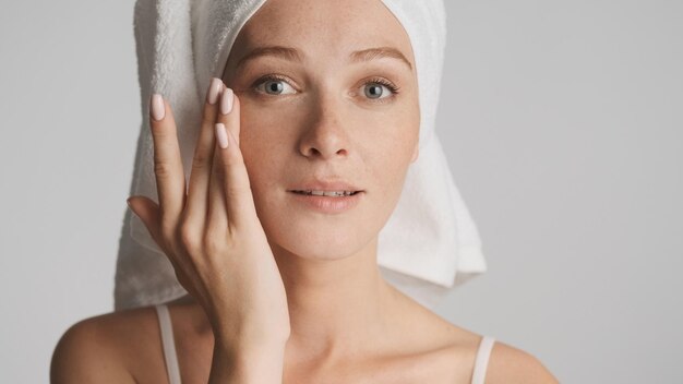 Крупным планом молодая женщина с гладкой кожей и полотенцем на голове чувственно смотрит в камеру на белом фоне