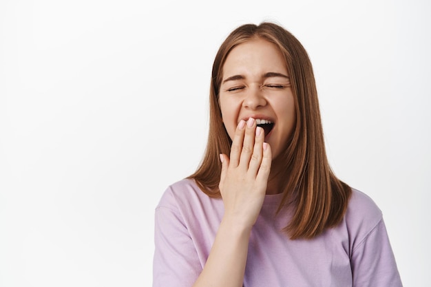 Primo piano di una giovane donna che sbadiglia, studente stanco durante gli esami, che si sveglia presto, che copre la bocca aperta annoiato, in piedi esausto su sfondo bianco